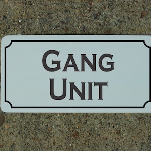 May include: White rectangular sign with a black border and rounded corners. The sign reads "GANG UNIT" in black serif font.