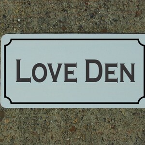 May include: White rectangular sign with a black border and rounded corners. The sign says "LOVE DEN" in black serif font.