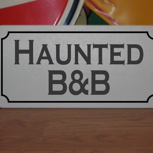 Puede incluir: Un letrero blanco con letras negras que dice "HAUNTED B&B". El letrero tiene un borde decorativo.