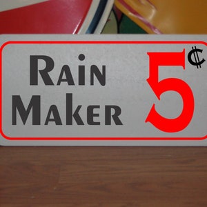 May include: A white rectangular sign with red trim and black text that reads "Rain Maker 5¢". The number 5 is red and the cent symbol is black.