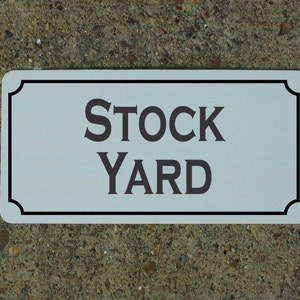 May include: White rectangular sign with a black border and rounded corners. The sign says "STOCK YARD" in black lettering.