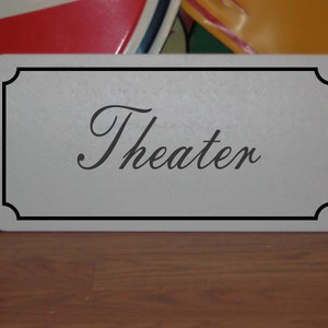 May include: A white sign with a black border and the word "Theater" written in a cursive font.