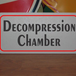 May include: White sign with red border, reading "Decompression Chamber" in black bold letters.