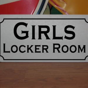 Puede incluir: Señal blanca con letras negras que dice "GIRLS LOCKER ROOM".