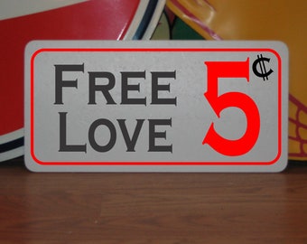 May include: A white sign with red lettering that says "FREE LOVE 5¢". The sign is on a wooden surface.