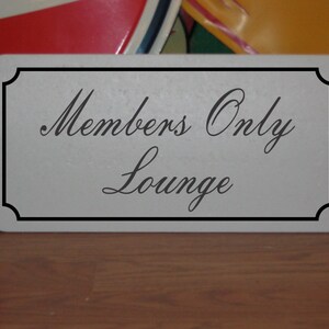 Peut inclure: Une pancarte blanche avec une bordure noire et le texte "Members Only Lounge" en lettres cursives noires.