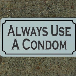 Może przedstawiać: Prostokątny znak z zaokrąglonymi rogami, z napisem "ALWAYS USE A CONDOM" w ciemnoszarych literach. Znak ma jasnoniebieskie tło z czarną obwódką, na teksturowanej szarej powierzchni.