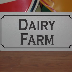 May include: A white metal sign with black trim and the text "DAIRY FARM" in black lettering.