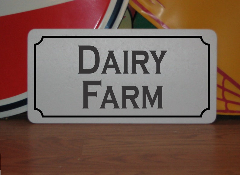 May include: A white metal sign with black trim and the text "DAIRY FARM" in black lettering.