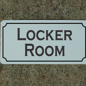 May include: A rectangular sign with rounded corners, featuring the words "LOCKER ROOM" in dark gray capital letters. The sign has a light blue background and a black border. The sign is on a textured gray surface.