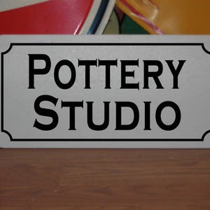 May include: A white sign with black lettering that reads "Pottery Studio".