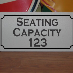 Op de afbeelding: Een wit rechthoekig bord met een zwarte rand en afgeronde hoeken. Het bord geeft "ZITPLAATSEN 123" aan.