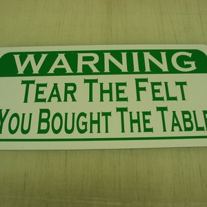 May include: White rectangular sign with green border and text that reads "WARNING TEAR THE FELT YOU BOUGHT THE TABLE"