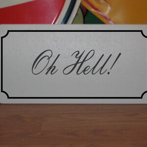 Puede incluir: Un letrero blanco con borde negro y el texto "Oh Hell!" en letras cursivas negras.