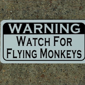 May include: White sign with black text that reads "WARNING WATCH FOR FLYING MONKEYS".