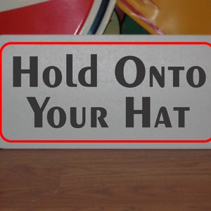 May include: White sign with a red border, reading "Hold Onto Your Hat" in black bold letters.