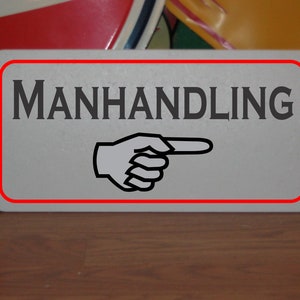 Peut inclure: Un panneau blanc avec une bordure rouge qui dit "MANHANDLING" en lettres noires. Une main pointant noire et blanche est en dessous du texte.