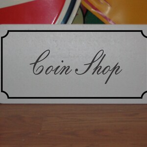 May include: A rectangular sign with a white background and a black border. The words "Coin Shop" are written in elegant cursive script. The sign is placed on a wooden surface.