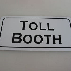 May include: White rectangular sign with black text that reads "TOLL BOOTH".