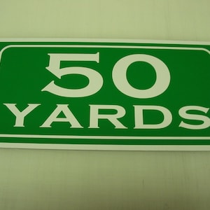 May include: A green rectangular sign with a white border and white text that reads "50 YARDS".