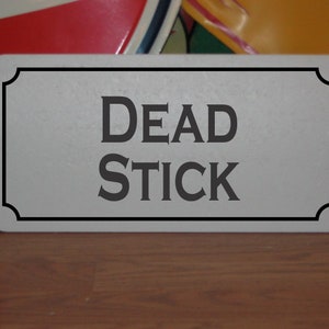 May include: A white rectangular sign with a black border and rounded corners. The sign has the text "DEAD STICK" in black lettering.
