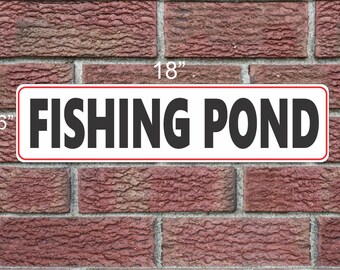 Puede incluir: Un letrero rectangular con la inscripción "FISHING POND" en letras negras y en negrita. El letrero mide 45,7 cm de largo y 15,2 cm de alto, con fondo blanco y borde rojo. Está montado sobre una pared de ladrillos rojos.
