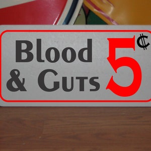 May include: A white sign with a red border and black text that reads "Blood & Guts 5¢". The number 5 is red and the cent symbol is black.