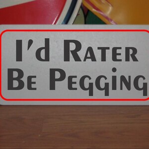 May include: A rectangular sign with a red border and the text "I'd Rater Be Pegging" in dark gray letters. The sign is on a wooden surface, with blurred objects in the background.