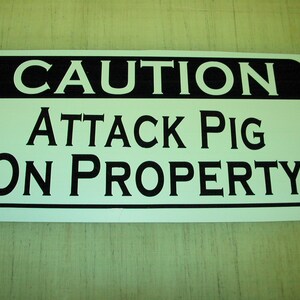 Könnte beinhalten: Ein weißes Schild mit schwarzem Text, auf dem steht "ACHTUNG ANGRIFFSSCHWEIN AUF GRUNDSTÜCK".
