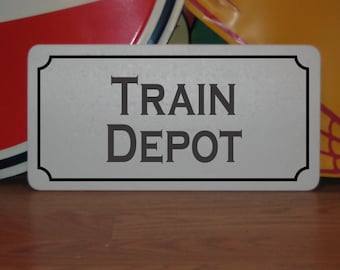 Può includere: Un cartello rettangolare con le parole "TRAIN DEPOT" in grigio scuro, centrato su uno sfondo grigio chiaro. Il cartello ha un bordo nero con angoli arrotondati. Il cartello è su una superficie di legno.