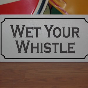 May include: A white rectangular sign with black lettering that reads "WET YOUR WHISTLE".