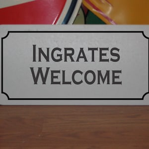 May include: A white sign with black lettering that reads "Ingrates Welcome". The sign has a decorative border.