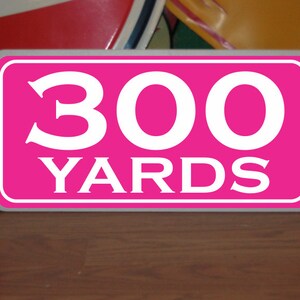 May include: A pink rectangular sign with white lettering that reads "300 YARDS".