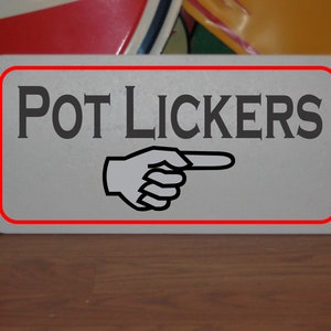 Puede incluir: Un letrero rectangular con las palabras "POT LICKERS" en letras grises oscuras. Una mano con un dedo señalando está debajo del texto. El letrero tiene un borde rojo y está sobre una superficie de madera.