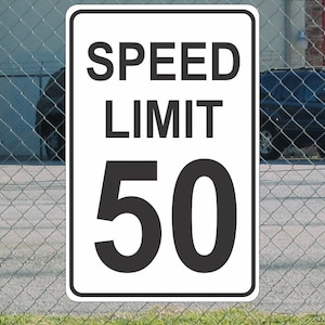 Op de afbeelding: Een rechthoekig wit snelheidslimietbord met zwarte tekst en cijfers. Het bord luidt "SPEED LIMIT 50". Het bord is gemonteerd op een hek van gaas met een vage achtergrond van een gebouw en een auto.