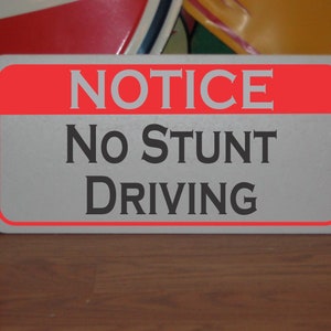 May include: A rectangular sign with a red border and a red top section that reads "NOTICE" in gray letters. Below, the sign says "No STUNT DRIVING" in a bold, gray font. The sign is on a wooden surface.