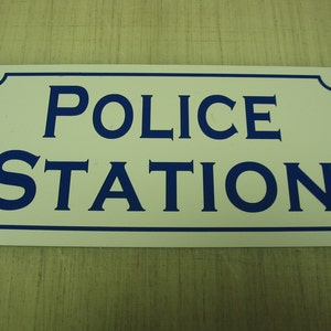 May include: White rectangular sign with blue lettering that reads "POLICE STATION".