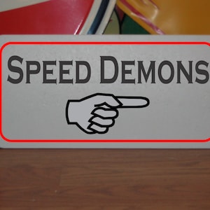 Puede incluir: Un letrero rectangular con borde rojo muestra las palabras "SPEED DEMONS" en gris oscuro, sobre una mano que señala. El letrero es gris claro y parece ser de metal. El fondo está borroso.