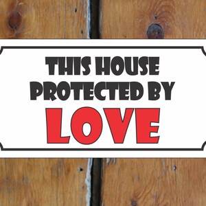 Puede incluir: Un cartel blanco con borde negro y esquinas redondeadas. El cartel dice "This house protected by LOVE" en letras negras y rojas.