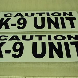 May include: Two white signs with black text that reads "CAUTION K-9 UNIT".