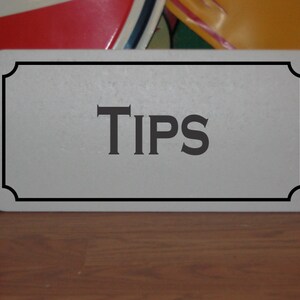 May include: A white rectangular sign with a black border and rounded corners. The sign has the word "TIPS" printed in black letters.