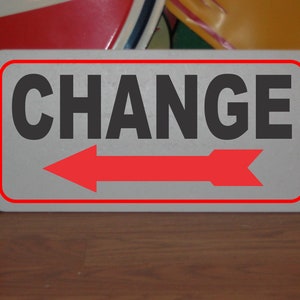 May include: A white sign with a red border and a red arrow pointing to the left. The sign says "CHANGE" in black letters.