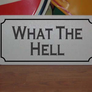 May include: A white rectangular sign with black trim and rounded corners. The sign has the text "WHAT THE HELL" in black lettering.
