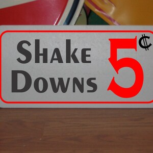 Könnte beinhalten: Ein weißes Schild mit rotem Rand und schwarzem Text, auf dem "Shake Downs 5¢" steht. Die Zahl 5 ist rot und das Cent-Symbol ist schwarz.