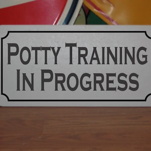 May include: White sign with black text that reads "Potty Training In Progress".