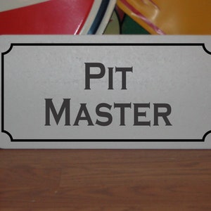 May include: A white rectangular sign with a black border and rounded corners. The sign reads "PIT MASTER" in black serif font.