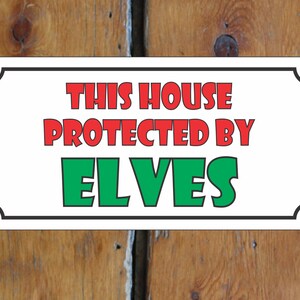 Może przedstawiać: Biały znak z czarną ramką i zaokrąglonymi rogami. Znak zawiera napis "This house protected by elves" czerwonymi i zielonymi literami.