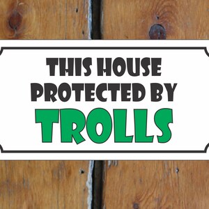 Puede incluir: Un cartel blanco con borde negro y esquinas redondeadas. El cartel dice "This house protected by trolls" en letras negras y verdes.