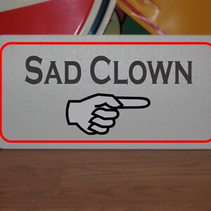 May include: A white sign with a red border and the text "SAD CLOWN" in black. A black and white pointing hand is below the text.