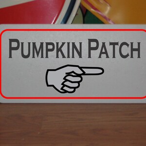 May include: A white sign with a red border and a black pointing hand. The sign says "Pumpkin Patch" in black letters.